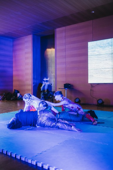 Four performers in costume on low stage, lit in blue