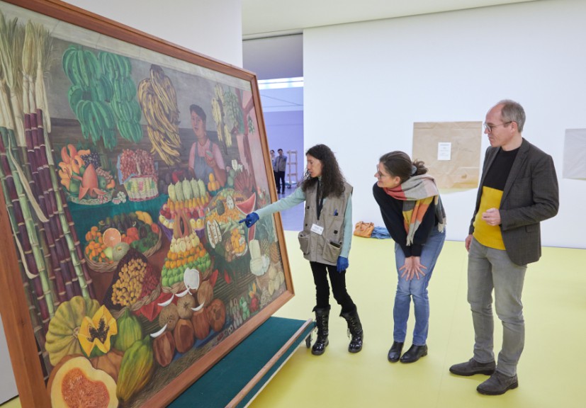 Two women and a man look at a large colourful painting up close